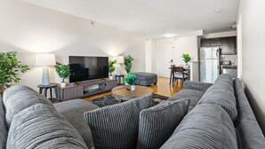Moderni olohuone, jossa on suuri harmaa sohva, litteänäyttöinen televisio sekä ruokailutila, jossa on pöytä ja tuolit.