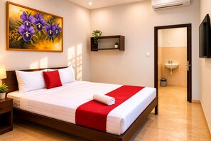 A hotel room with a bed, a painting of flowers, a bedside table with a lamp, a small shelf with a plant, and a bathroom visible through an open door. - Cahaya Adrian Near Komodo Airport (Labuan Bajo)