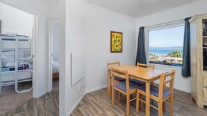 A room with a wooden table and chairs, a window with a view of the beach, a bed with a white headboard, and a white radiator. - Kuaotunu Vista - Ocean Views (Kuaotunu)