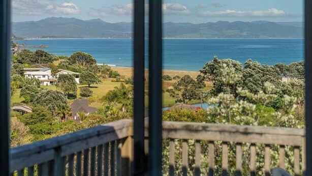 A coastal view from a balcony with a wooden railing, overlooking a beach, greenery, and distant mountains. - Kuaotunu Vista - Ocean Views (Kuaotunu)