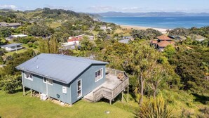 A coastal house with a deck, surrounded by lush greenery and a view of the beach and ocean. - Kuaotunu Vista - Ocean Views (Kuaotunu)