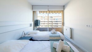 A hotel room with two beds, a large window with blinds, a radiator, and a view of a building outside. - 2-Br Panoramic Sea View & Beach Access (Cannes)