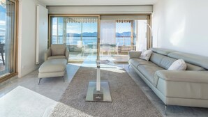 A modern living room with a sofa, armchairs, a glass coffee table, and a view of the sea through large windows. - 2-Br Panoramic Sea View & Beach Access (Cannes)