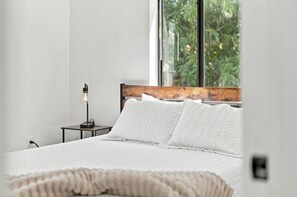 A bedroom with a bed, a nightstand, a lamp, and a window with a view of greenery.