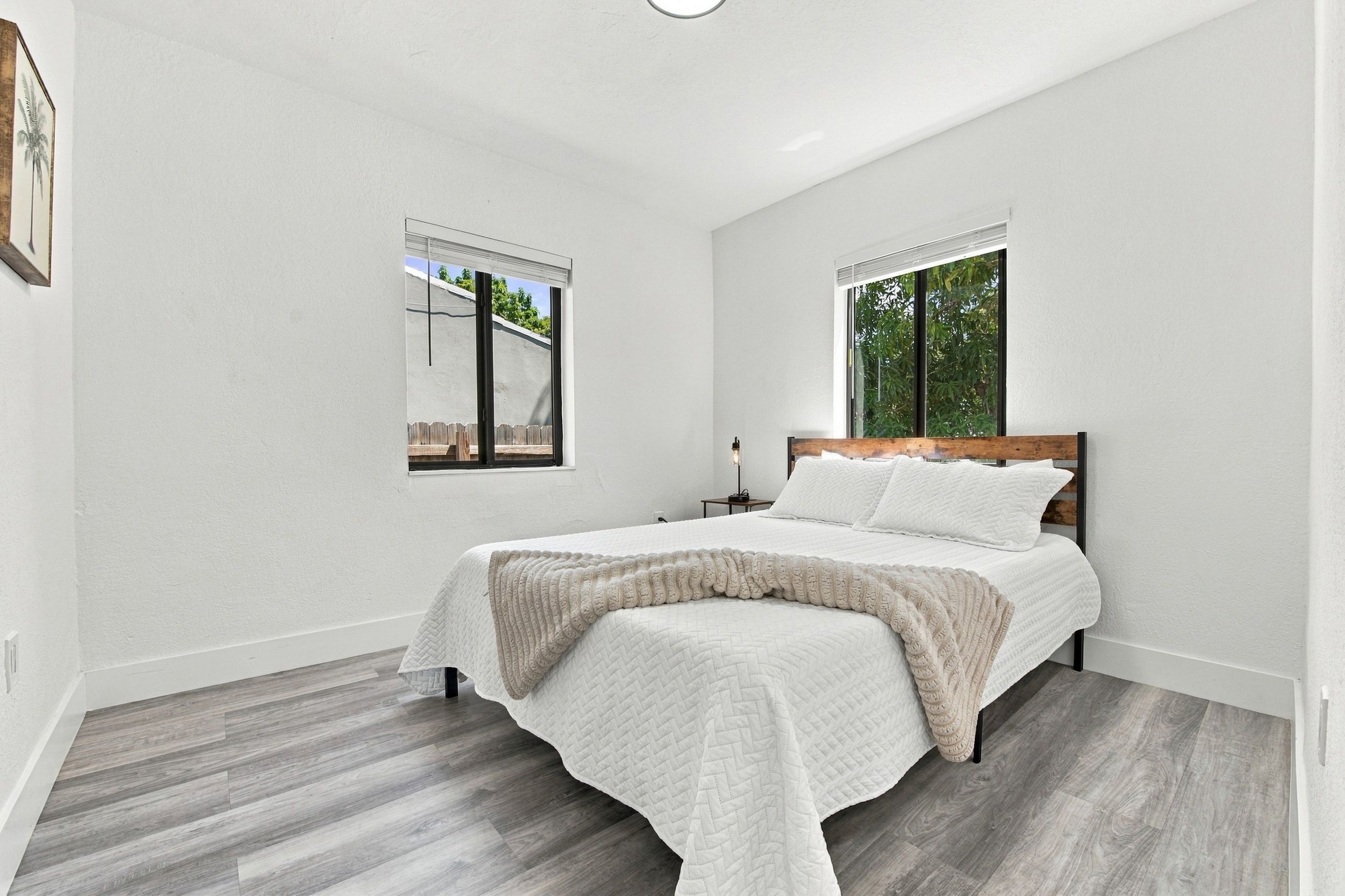 A bedroom with a bed, two windows, a nightstand, and a view of the outdoors.