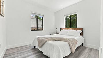 A bedroom with a bed, two windows, a nightstand, and a view of the outdoors.