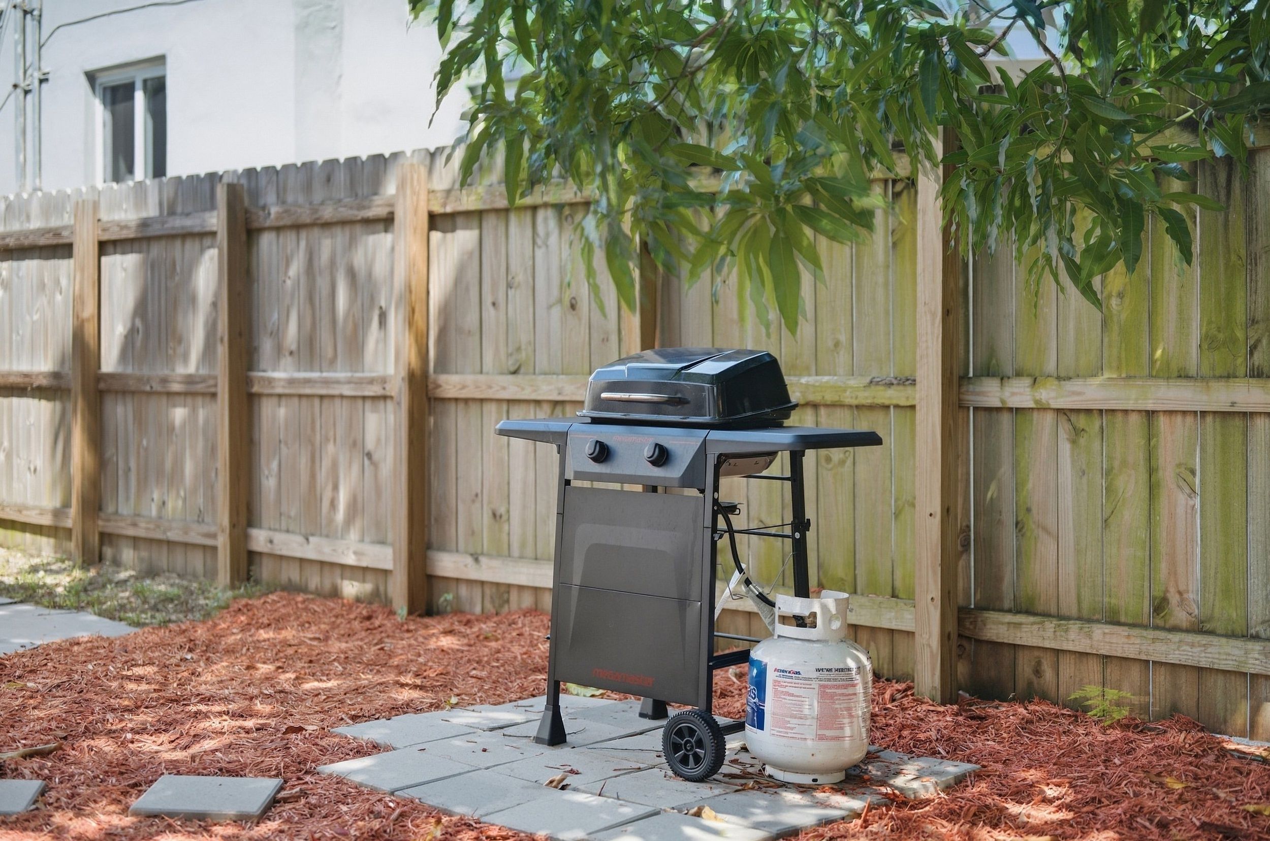 A portable barbecue grill with a propane tank in a backyard with a wooden fence and mulch.