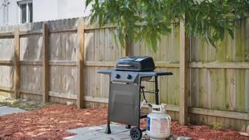 A portable barbecue grill with a propane tank in a backyard with a wooden fence and mulch.