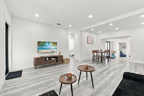A spacious living area with a flat-screen TV, a wooden entertainment unit, and a dining area with a table and chairs.