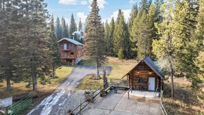 Une cabane dotée d’un porche et d’un bâtiment séparé comprenant un garage, le tout entouré d’une forêt dense.
