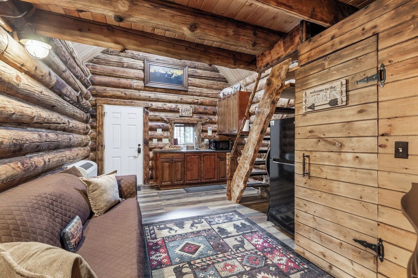 A log cabin interior with a cozy living area, a kitchen with wooden cabinets, and a staircase leading to another level.