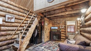 L’intérieur d’une cabane en rondins, avec un escalier en bois, une cheminée, un téléviseur et un coin salon confortable.