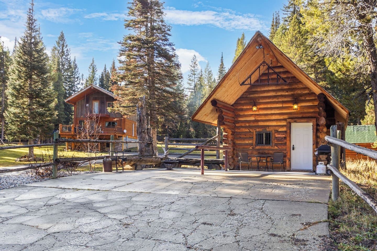 A rustic log cabin with a porch and outdoor seating area, surrounded by tall trees.