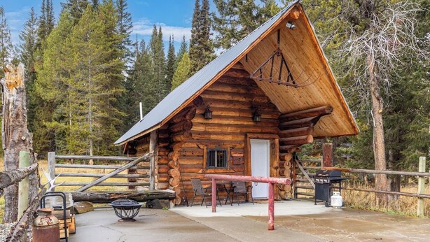 Une cabane rustique en rondins de bois, avec un toit en métal, une véranda et un espace aménagé pour faire un feu de camp.