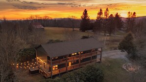 A two-story log cabin with multiple balconies and a deck, surrounded by trees and a grassy field. - Designer | Hot Tub | Gameroom | Firepit | Sleeps12 (Banner Elk)