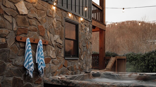 A stone exterior wall with a hot tub, wooden balcony, and string lights. - Designer | Hot Tub | Gameroom | Firepit | Sleeps12 (Banner Elk)