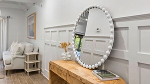 Una habitación con un banco de madera, un espejo con marco blanco, un jarrón con flores secas y un libro sobre el banco.