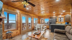 A wooden cabin interior with a dining area, kitchen, and a large window offering a view. - 3BR | Long Range Views | Hot Tub | Minutes to Pkwy (Sevierville)