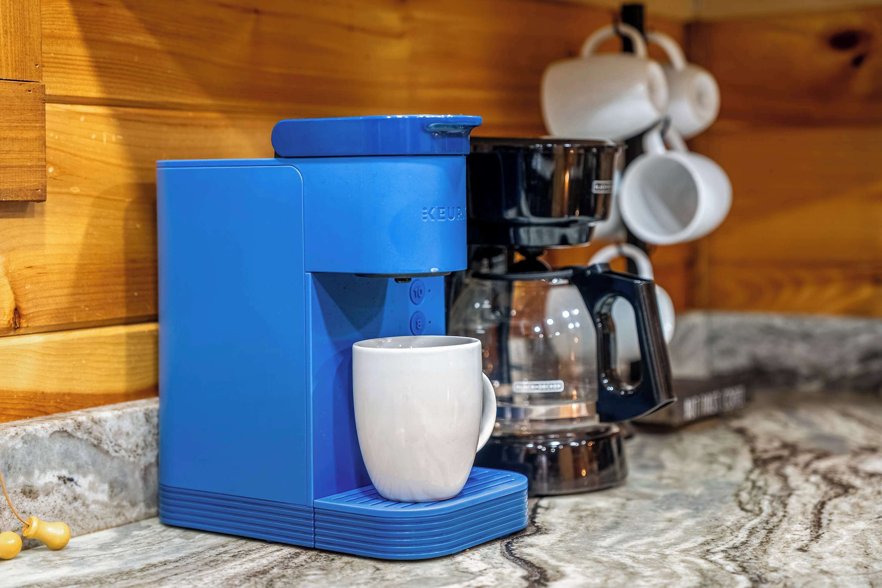A blue Keurig coffee maker with a white mug on a coaster, a glass carafe, and a coffee pot on a wooden wall.