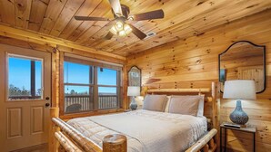 A wooden cabin bedroom with a bed, ceiling fan, windows, and a nightstand. - 3BR | Long Range Views | Hot Tub | Minutes to Pkwy (Sevierville)