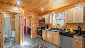 Interior de una cabaña de madera con cocina, una puerta roja, lavadora y ventana con vistas.