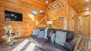 Interior de una cabaña de madera con un sofá de cuero, un televisor y una escalera.