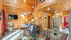 Interior de una cabaña de madera con sofá, televisor, escalera y comedor.