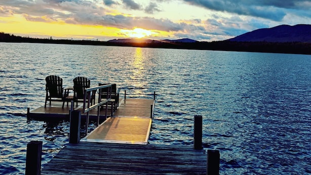 Une jetée en bois s’étendant sur une étendue d’eau calme, agrémentée de chaises et d’une table, le tout entouré d’un paysage montagneux et illuminé par un coucher de soleil.