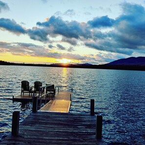 Een houten steiger die uitloopt in kalm water, met stoelen en een tafel, omgeven door een bergachtig landschap en een zonsondergang.