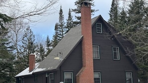 Une maison à deux étages, dotée d’une cheminée en briques imposante, entourée de neige et d’arbres.