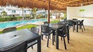 An outdoor dining area with a pool, covered by a thatched roof, and a view of a modern building in the background.