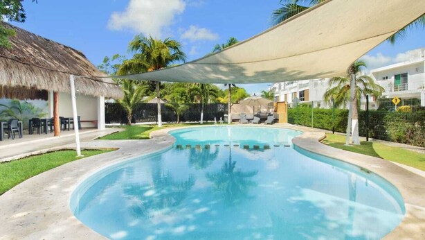 A swimming pool with a thatched roof structure, palm trees, and a white building in the background.