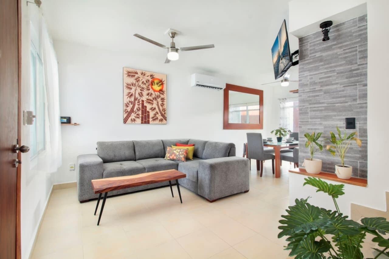 Un salon moderne avec un canapé gris, une table basse en bois, un ventilateur de plafond et un coin repas comprenant des chaises et une table.