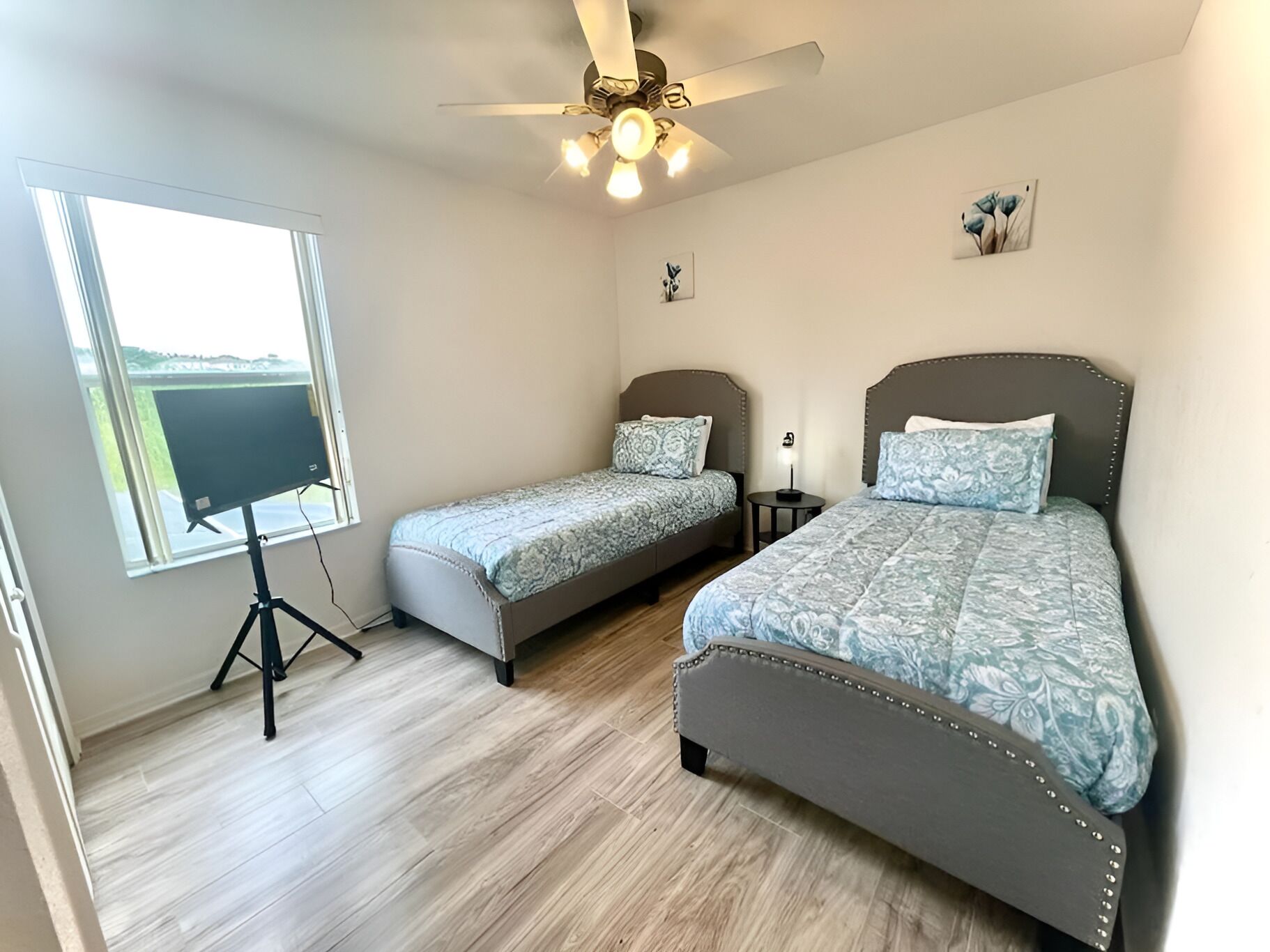 Une chambre avec deux lits, un ventilateur de plafond, une lampe sur pied et une fenêtre donnant sur un beau paysage.