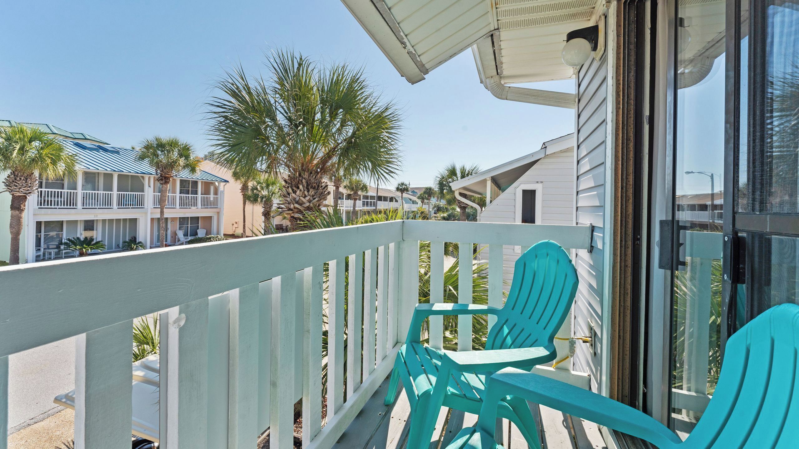 A balcony with turquoise chairs, a white railing, and a view of a resort building with multiple floors and palm trees.