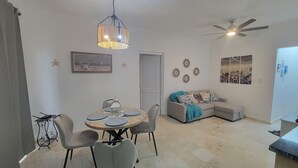 Uma sala de jantar com uma mesa redonda, cadeiras e um lustre. Há um sofá com manta e almofadas decorativas ao fundo.