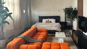 A modern bedroom with a large orange sectional sofa, a bed with a black headboard, a TV, and a potted plant. - Detroit Studio Loft Cozc Private Entry (Detroit)