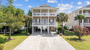 A two-story building with a central entrance, a covered porch, and a small garden area with palm trees and flowering plants. - Spacious 5-bedroom house with pool near ocean and amenities in Murrells Inlet (Murrells Inlet)
