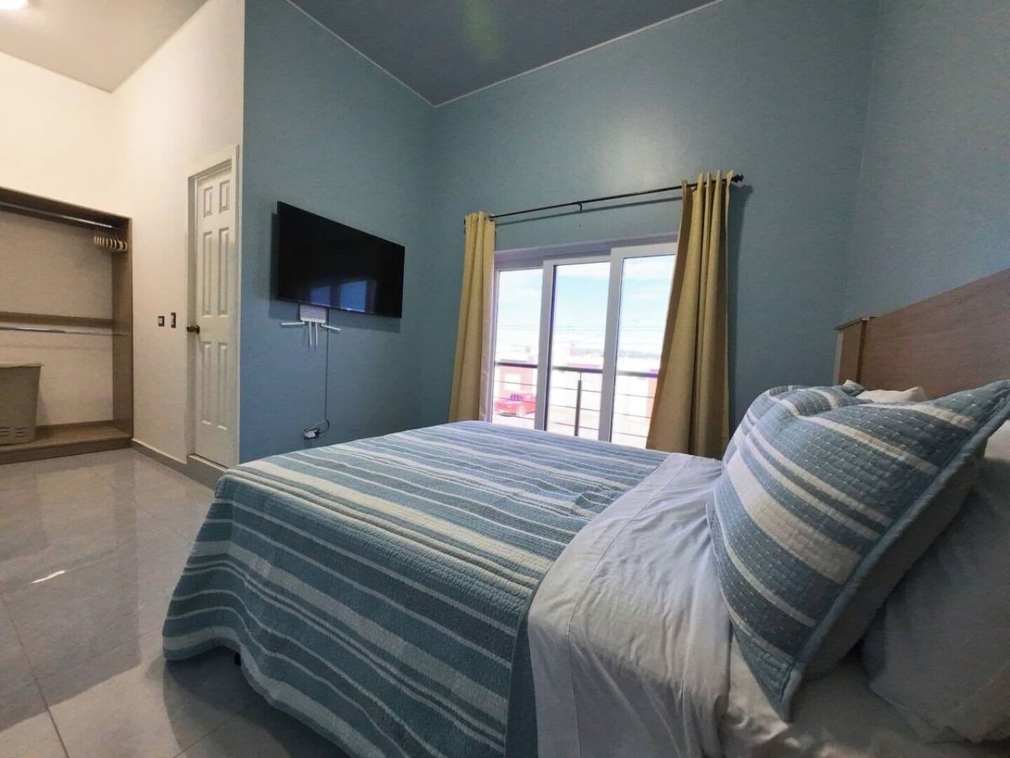 Un dormitorio con cama, televisor montado en la pared, ventana con cortinas y vistas al balcón.