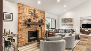 A living room with a stone fireplace, grey sofas, a flat-screen TV, and a coffee table. - Design-forward Palm Springs Home w/ Saltwater Pool, Lush Yard & Multiple Outdoor Dining Areas Casa Papagayo by Avantstay (Palm Springs)