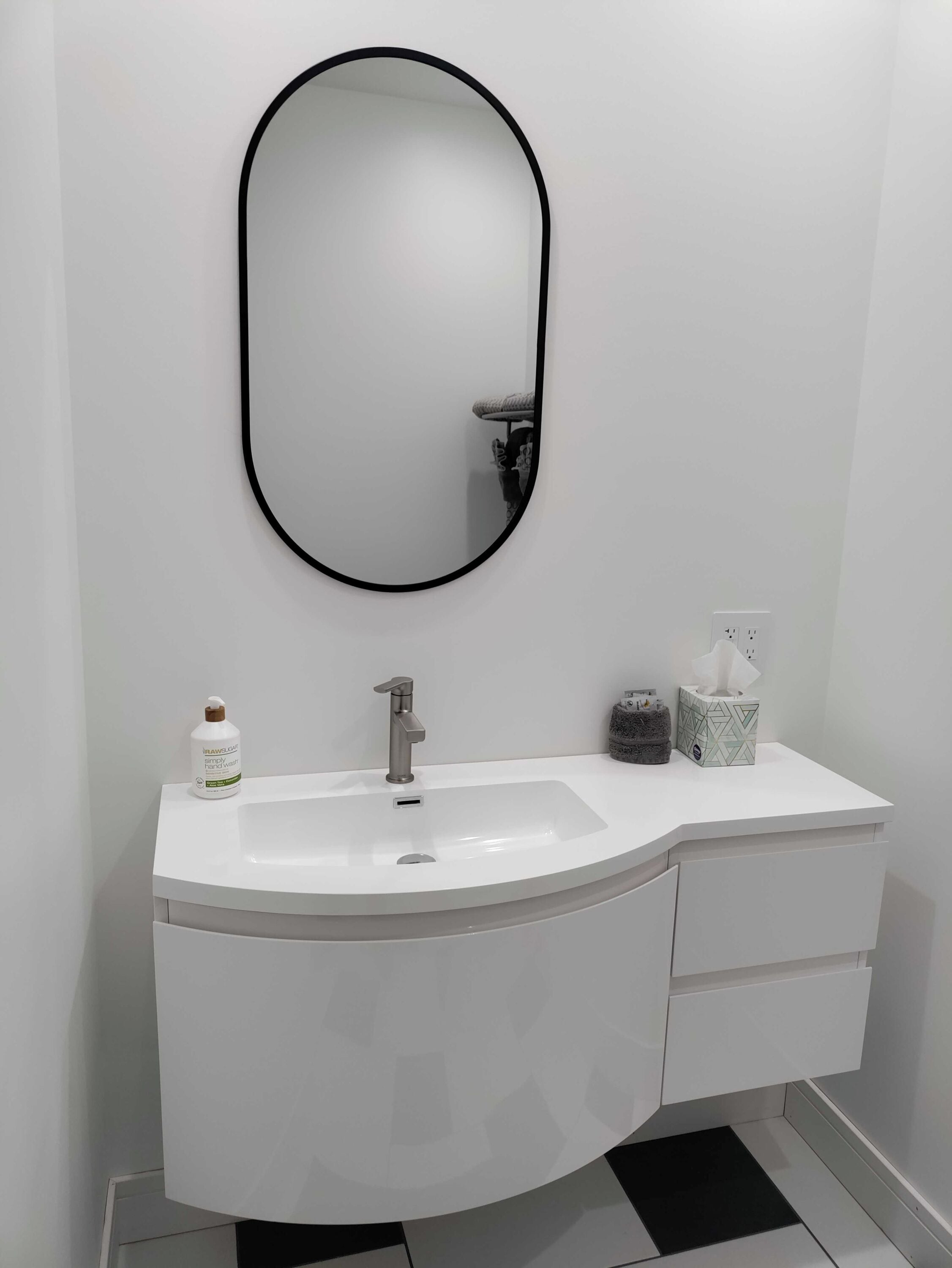 A modern bathroom with a white vanity, a round mirror with a black frame, a silver faucet, and a small bottle of hand soap.