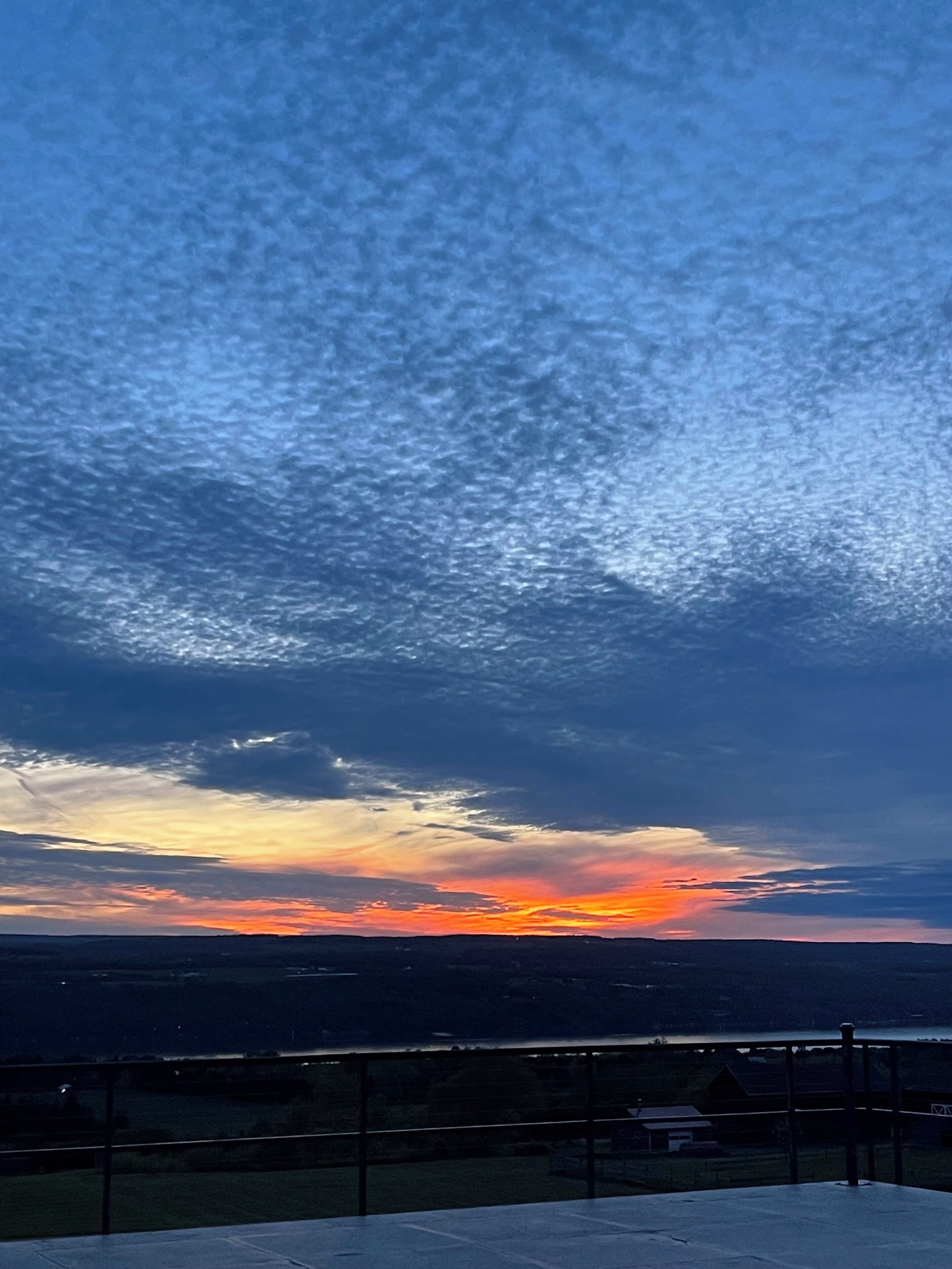 A sunset view from a balcony with a railing, overlooking a landscape with distant buildings.