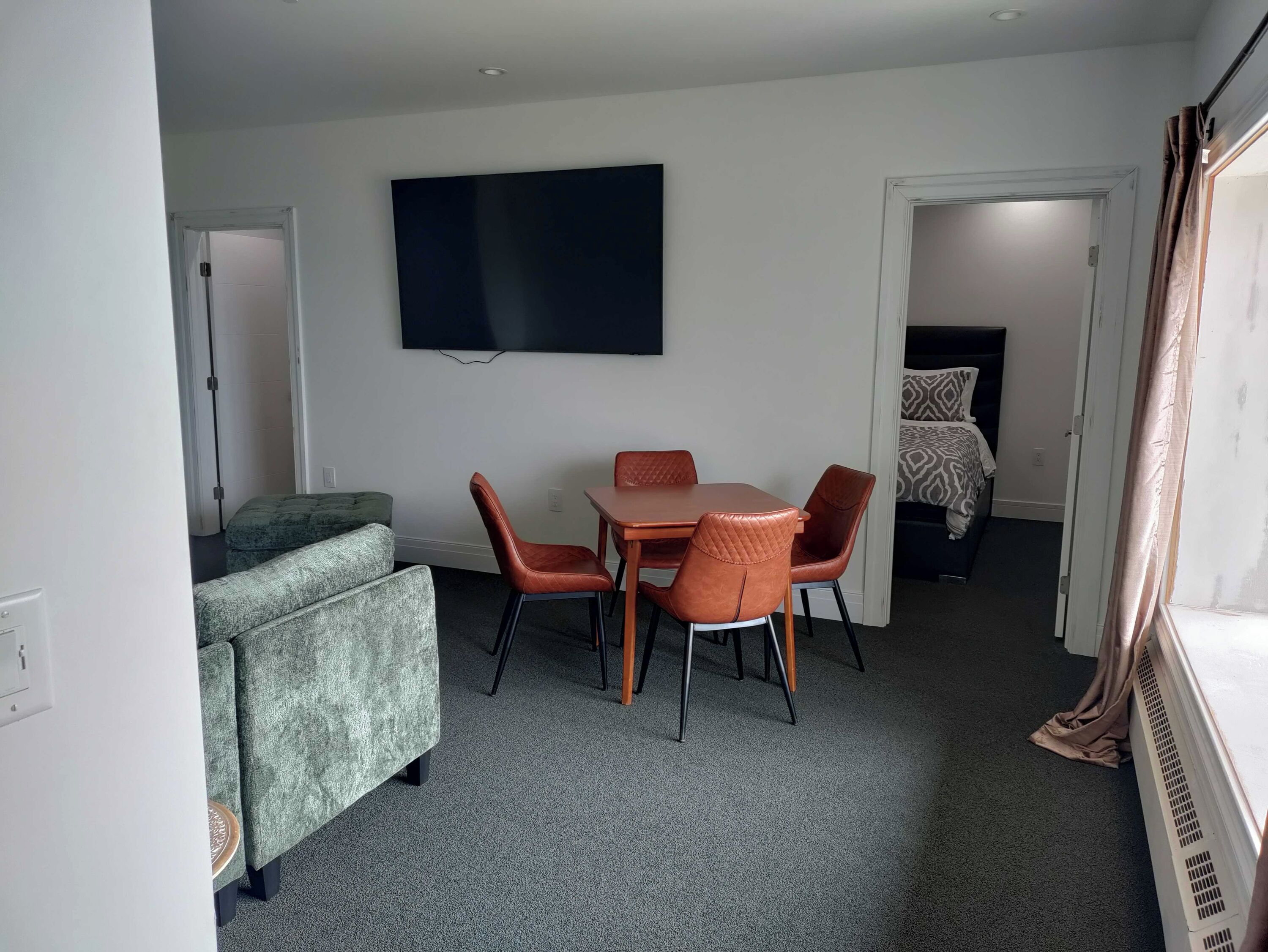 A compact room with a grey carpet, a small table with four chairs, a green sofa, a flat-screen TV mounted on the wall, and a bed with patterned bedding.
