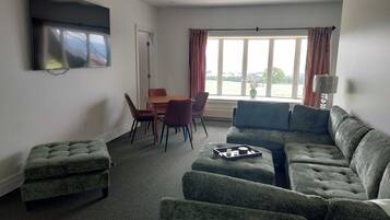 A living room with a grey sofa, a dining table with chairs, a flat-screen TV mounted on the wall, and a window with curtains.