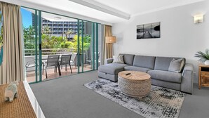 A modern living room with a grey sofa, a round wooden coffee table, and a large glass door leading to an outdoor balcony with a view of a building and greenery. - Sunlit 1-Bedroom Apt at Crown Tower with Parking (Surfers Paradise)