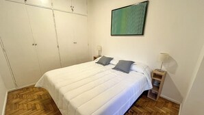 Un dormitorio con una cama, dos almohadas, una mesita de noche, una lámpara y un armario.