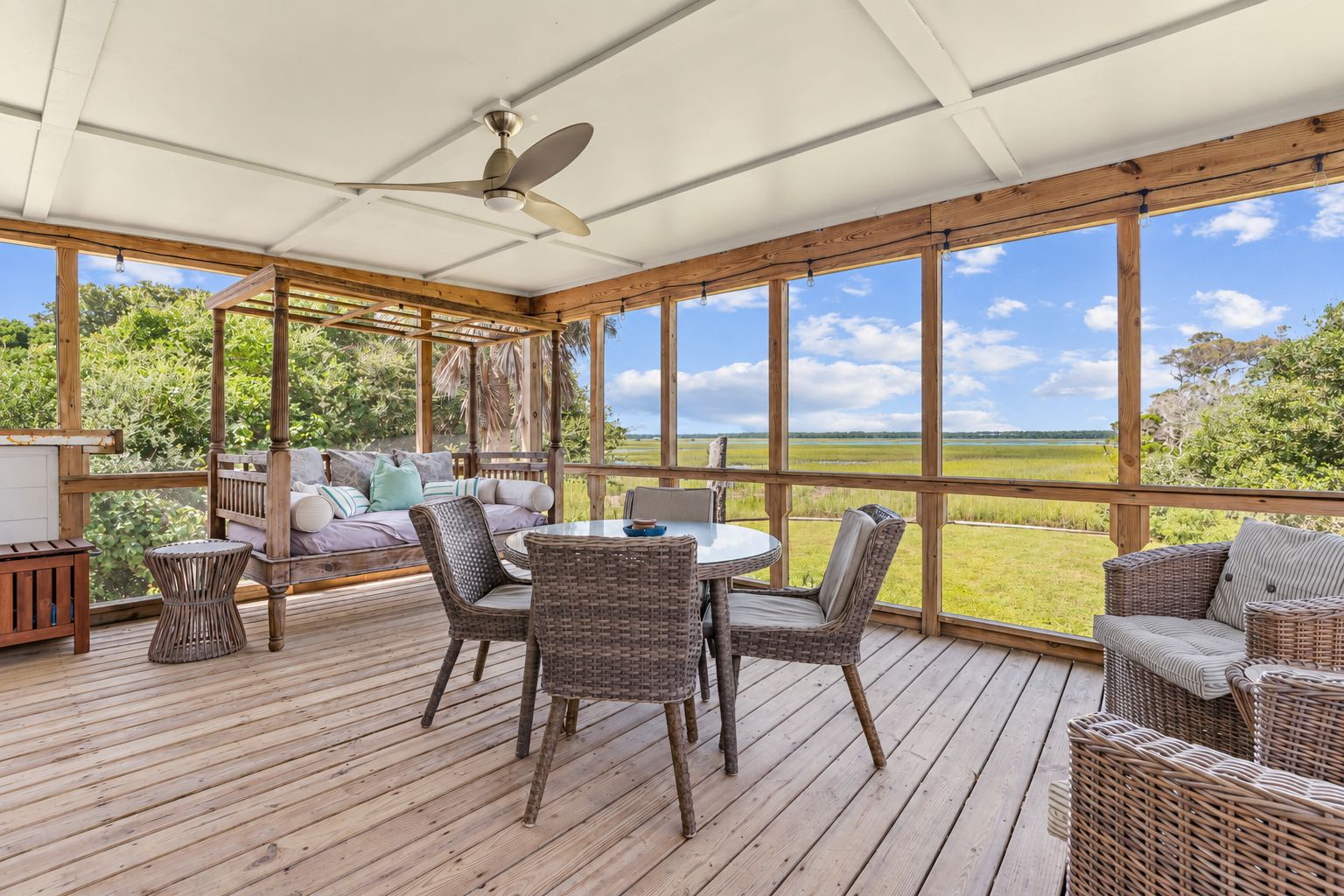 A covered outdoor area with a wooden deck, wicker furniture, and a view of a grassy field.