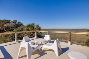 Una terraza con sillas de plástico blanco y una mesa, con vistas a una playa y una zona pantanosa.