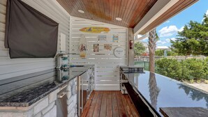 An outdoor kitchen area with a wooden ceiling, a stone wall, and a glass enclosure. - HEATED POOL. Backyard retreat! Game room. 3-min walk to beach via backyard path (Surf City)
