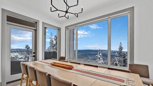 A modern dining room with a long table, multiple chairs, and large windows offering a snowy mountain view. - Western Star Chalet- Ski-in/out & Amazing Mtn View (Big White)